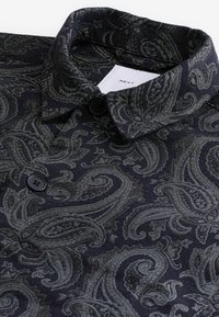 Chemise bleu foncé avec motif cachemire argenté, col boutonné et deux boutons noirs visibles, avec une étiquette blanche portant le nom de la marque NEXT.