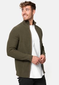 Suéter de punto verde oliva con cremallera, textura acanalada, cuello alto y puños ajustados. Llevado sobre una camiseta blanca, diseño de corte ajustado.
