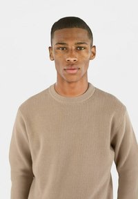 Beige ribbestrikket genser med rund hals, som har lange ermer og en teksturert overflate. Stoffet ser mykt og behagelig ut.