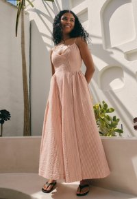 Robe rayée longue, rose et blanche avec un corsage ajusté et une jupe fluide. Présente des bretelles fines et un tissu texturé. Portée avec des sandales noires.