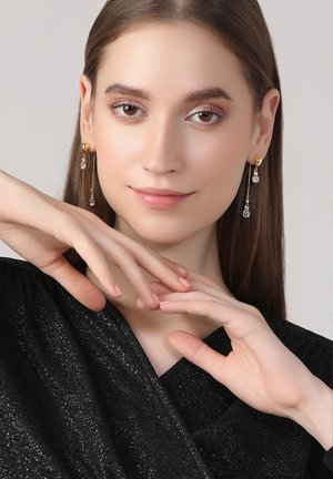 Gold- und Silber-Ohrringe mit klaren Kristallen, die ein elegantes Design aufweisen. Das Model trägt ein strukturiertes schwarzes Oberteil und zeigt die Hände anmutig.