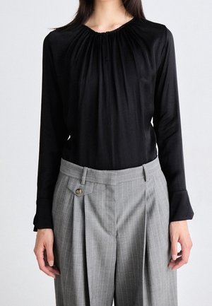 Personne portant un chemisier noir à manches longues avec encolure froncée et un pantalon gris à rayures fines taille haute, faisant face à un fond uni.