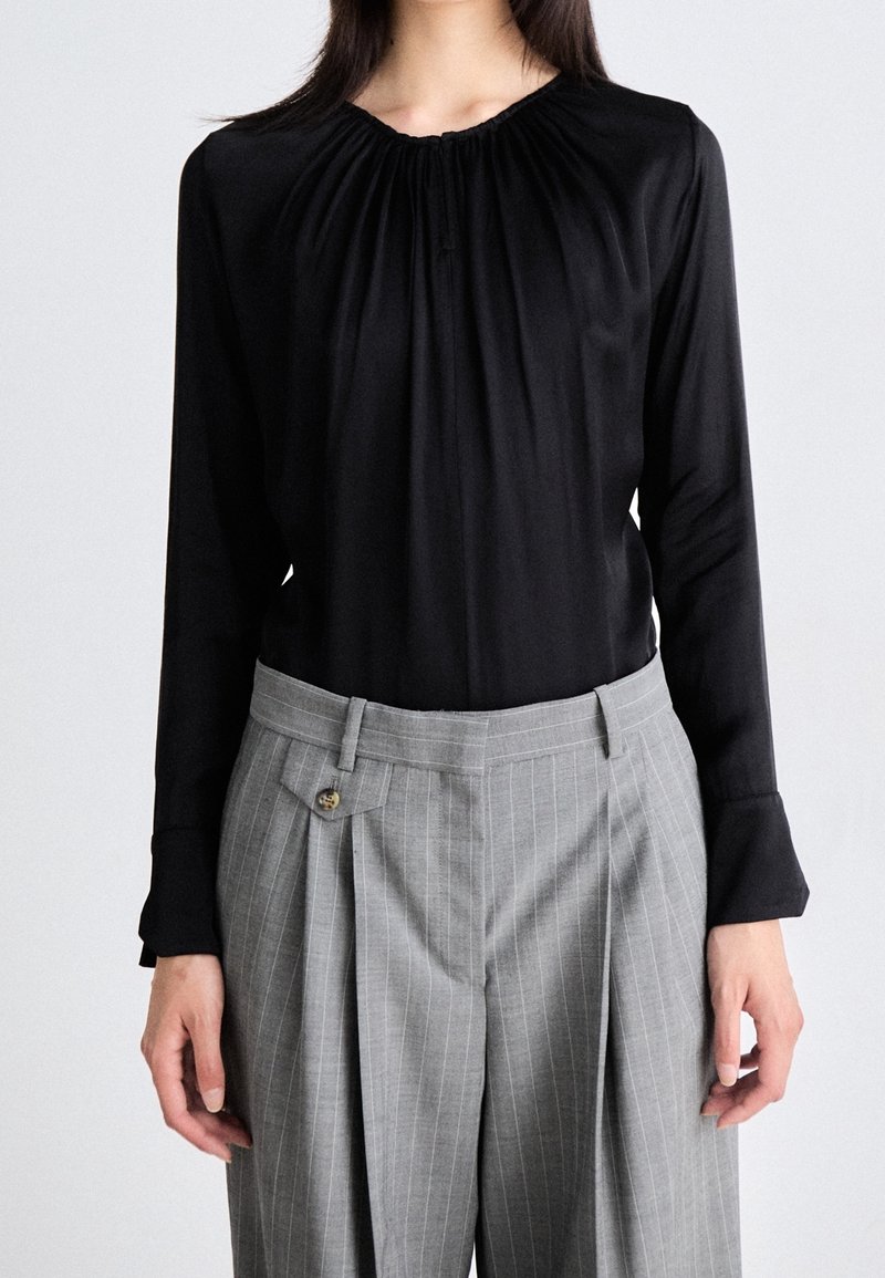 Personne portant un chemisier noir à manches longues avec encolure froncée et un pantalon gris à rayures fines taille haute, faisant face à un fond uni.