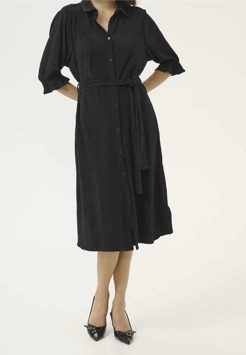Femme portant une robe chemise midi texturée noire avec une ceinture à la taille et une fermeture à boutons, associée à des talons noirs à bout pointu.