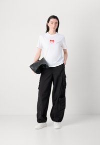 Witte t-shirt met een rood logo, gecombineerd met zwarte cargobroeken met zakken en ritsen, en witte sneakers. Het model houdt een zwarte clutch vast.