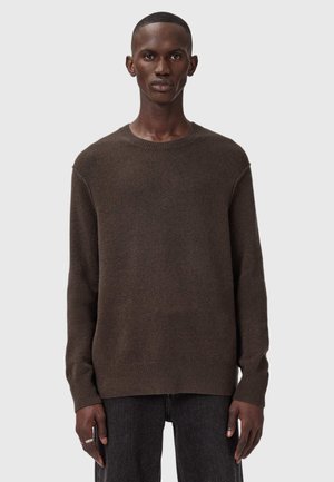 Man wearing a dark brown long-sleeve crew-neck sweater and black pants, standing against a plain light gray background.