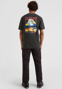 T-shirt in cotone grigio scuro con una grafica colorata sulla schiena, comprensivo di testo e illustrazioni, abbinato a pantaloni e scarpe neri.