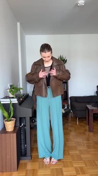 Personne ajustant une veste en cuir marron par-dessus un haut noir et un pantalon large bleu clair, debout dans un salon confortable avec des plantes et des meubles.