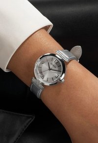 Hõbedast kell, millel on ümar dial, hall nahkrihm ja kristallaktsendid kella hoobadel. Omab suuri numbreid ja tekstureeritud nägu.