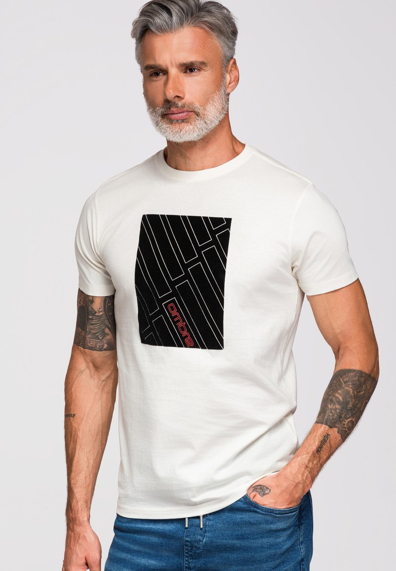 Homme d'âge moyen aux cheveux gris et barbe, portant un T-shirt blanc avec un motif géométrique noir et un texte rouge, un jean bleu et des bras tatoués.