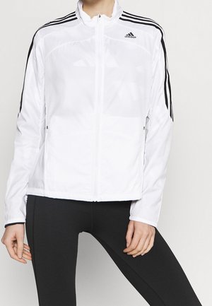 Person iført en hvid Adidas lynlåsjakke med sorte striber på ærmerne og sorte leggings, stående mod en ensfarvet baggrund.