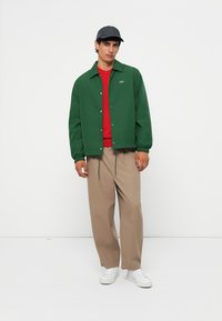 Chaqueta verde con botones a presión, suéter rojo debajo, pantalones beige y zapatillas blancas. Gorra gris completa el conjunto.