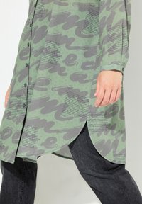 Groen button-up shirt met grijze abstracte patronen en tekst, lange mouwen, zijsplitsingen, gedragen over donkere jeans.