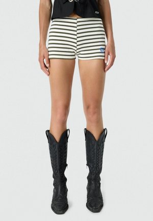 Persona con pantalones cortos blancos con rayas horizontales negras y botas de vaquero negras altas, posando contra un fondo liso.