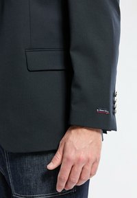 Blazer gris foncé à coupe ajustée, avec une poche poitrine, des boutons décoratifs sur la manchette, et une étiquette "Armor Lux" cousue sur la manche.