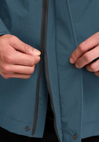 Lichte blauwe jas met een gestructureerd oppervlak en zwarte rits. Handen bedienen de rits en de drukknoopjes aan de voorkant.