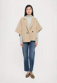 Beige double-breasted oversized coat with wide sleeves, worn over a gray turtleneck, paired with blue straight-leg jeans and leopard print slip-on shoes.