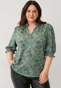 Grøn bluse med blomsterprint, v-hals, sammensunkne ærmer og en struktureret stof. Den har en afslappet pasform og bløde flæse detaljer.