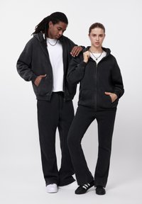 Μαύρα φούτερ με φερμουάρ και κουκούλα από βαμβάκι, με πλαϊνές τσέπες, συνδυασμένα με μαύρο παντελόνι σε άνετη γραμμή. Λεία υφή, απλός σχεδιασμός και ελάχιστα διακοσμητικά στοιχεία.