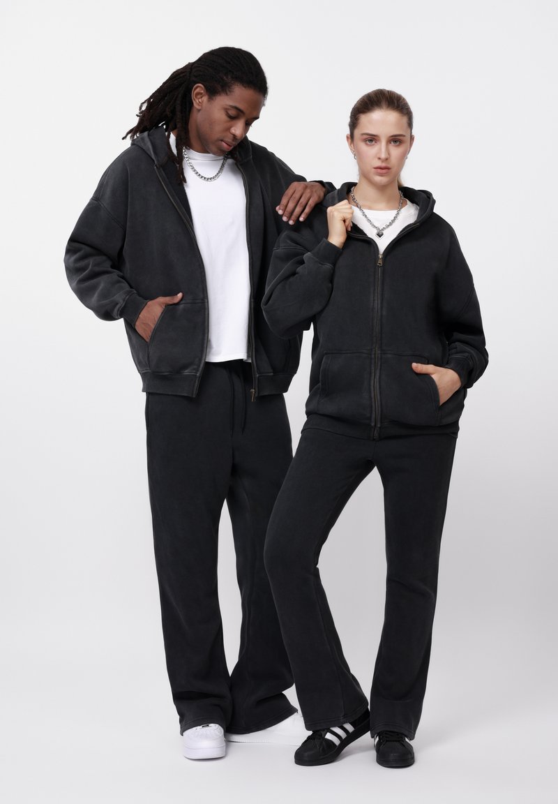 Μαύρα φούτερ με φερμουάρ και κουκούλα από βαμβάκι, με πλαϊνές τσέπες, συνδυασμένα με μαύρο παντελόνι σε άνετη γραμμή. Λεία υφή, απλός σχεδιασμός και ελάχιστα διακοσμητικά στοιχεία.