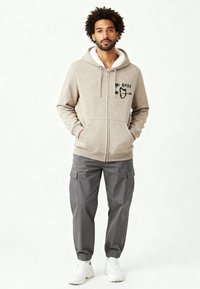 Sweat à capuche beige zippé avec une doublure en sherpa blanche, présentant un design noir brodé sur le devant. Porté avec un pantalon cargo gris et des baskets blanches.
