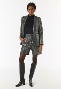 Błyszczący czarny żakiet z pasującymi szortami, oba z teksturowanym metalicznym wykończeniem; zestawione z czarnymi skórzanymi botkami do kolan.