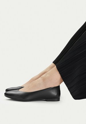 Czarne, skórzane baleriny z noskiem w szpic, noszone z czarnymi plisowanymi spodniami, widok z boku na jasnym tle.