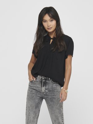 Zwarte blouse met korte mouwen en een sleutelgatsluiting, versierd met geplooide details, in combinatie met vervaagde grijze denim jeans. Eenvoudig, losvallend ontwerp.