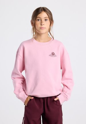 Jongeman in een roze sweatshirt met het "Golden Bagel" logo en bordeauxrode broek, staand met zijn handen in de zakken tegen een witte achtergrond.