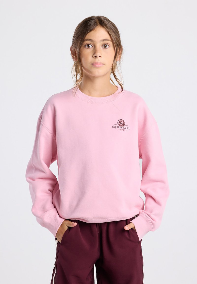 Personne jeune portant un sweat-shirt rose avec le logo "Golden Bagel" et un pantalon bordeaux, se tenant les mains dans les poches sur fond blanc.