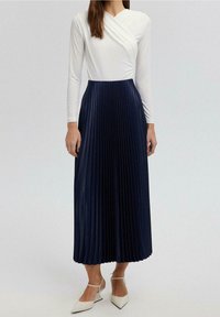 Top bianco a maniche lunghe, avvolgente, abbinato a una gonna maxi plissettata blu navy. La gonna presenta una texture verticale e raggiunge la caviglia.