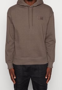 Felpa con cappuccio marrone realizzata in morbido misto cotone. Presenta una tasca frontale e una piccola etichetta con logo sul petto. Vestibilità leggermente più ampia.