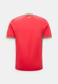 Κόκκινη αθλητική φανέλα με κοντά μανίκια, με πράσινη και λευκή διακοσμητική ρίγα στη λαιμόκοψη και τα μανίκια, αραβική γραφή τυπωμένη κάτω από τη λαιμόκοψη στην πλάτη.