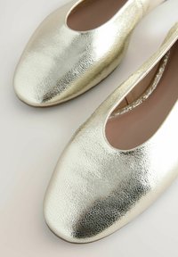 Metallisch goldene Ballerinas mit strukturierten Oberflächen, runder Spitze und einem schlupffreundlichen Design, das eine tiefe Öffnung an der Vorderseite aufweist.