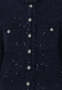 Marineblå paljetter cardigan med sølvknapper, med to brystlommer og et teksturert stoff med fletteDetaljer langs fronten.