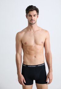 Homme torse nu avec des cheveux courts et foncés se tenant de face, portant un boxer noir Björn Borg sur fond clair.