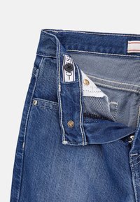 Blaue Jeans aus Denim mit Knopf- und Reißverschluss, ausgestattet mit einem weißen Innenfutter und kontrastierenden Nähten. Inklusive Logo-Patch und Nieten.