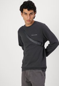 Sudadera gris oscuro de manga larga, con cuello redondo y un logo en la parte frontal. Presenta una correa diagonal en el pecho. Textura suave.
