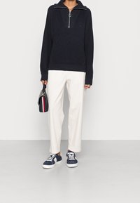 Pull en maille bleu marine avec un col zippé, pantalon large crème, et baskets bleues marines avec des accents blancs. Porte un petit sac à main noir.
