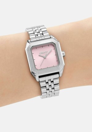 Montre en métal argenté avec un cadran carré rose affichant 10:08, portée sur un poignet avec un bracelet à maillons et une lunette rainurée.