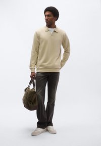 Világos bézs színű pulóver gallérral, sötét farmer, fehér sportcipő, zöld táska barna részletekkel, egyszínű háttér előtt állva.