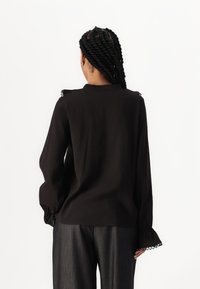 Blusa nera a maniche lunghe con scollo arricciato e polsini raccolti, dotata di rifiniture decorative a cerchio, abbinata a pantaloni grigi texture.