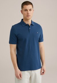 Temno modra polo majica s klasičnim ovratnikom, kratkimi rokavi in majhnim izvezenim logotipom na levem prsnem delu. Gladek, mehak material.