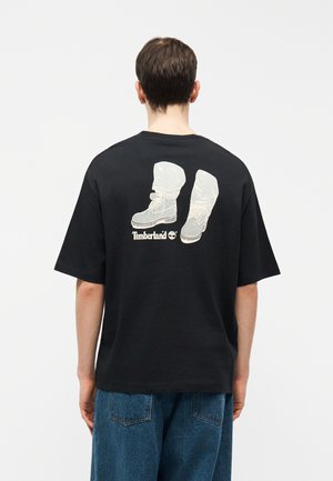 Osoba nosząca czarną koszulkę z białym nadrukiem butów i logo Timberland na plecach, w połączeniu z niebieskimi dżinsami.