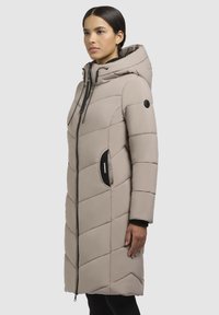 Langer, hellbeiger Puffermantel mit Kapuze und Chevron-Steppung. Verfügt über einen Reißverschluss vorne, seitliche Taschen und schwarze Akzente an Reißverschluss und Taschen.