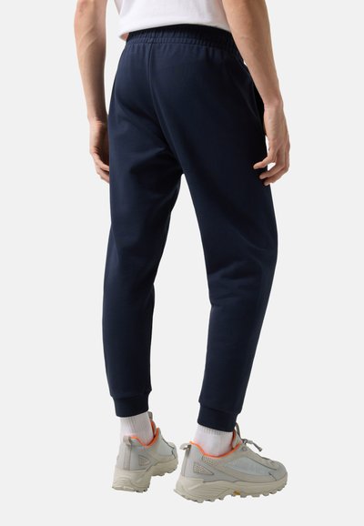 Mężczyzna w granatowych spodniach dresowych, białych skarpetkach i szarych trampkach z pomarańczowymi akcentami, stoi tyłem do kamery.