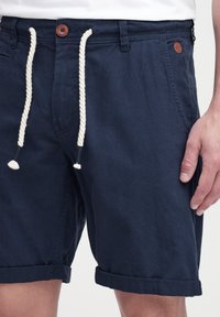 Pantalones cortos azul marino de algodón, con dobladillo enrollado, cintura con cordón en color blanco roto y una pequeña etiqueta de logo marrón en la parte delantera.
