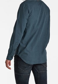 Camisa de manga larga en tejido verde oscuro con una ligera textura, presenta un dobladillo redondeado, puños abotonados y un detalle de costura en la parte posterior.