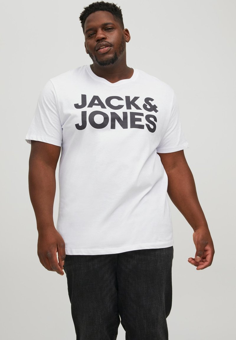 Weißes Baumwoll-T-Shirt mit großem, schwarzem "JACK & JONES"-Schriftzug auf der Vorderseite, kombiniert mit dunkelgrauen Jeans. Kurze Ärmel und Rundhalsausschnitt.