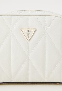Weiße gesteppte Tasche mit einem dreieckigen goldenen Logo, das "GUESS U.S.A." zeigt, akzentuiert durch ein texturiertes Muster und einen marineblauen Reißverschlussrand.
